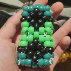 May include: A handmade beaded cuff bracelet in shades of green and black. The bracelet is constructed with small, round beads strung together with white string. The design features alternating rows of green, black, and teal beads, creating a geometric pattern.