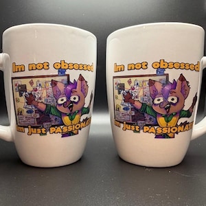 May include: Two white ceramic mugs featuring a cartoon character with purple hair standing in front of a corkboard with red string and papers. The text reads, "Im not obsessed, im just PASSIONATE" in orange and black lettering on the front of each mug.