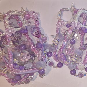 May include: Two decorative phone cases in shades of lavender. Each case is adorned with beads, flowers, and hanging purple orbs. The cases have a whimsical design, with a fairy-tale aesthetic. The cases are approximately 15 cm in length.