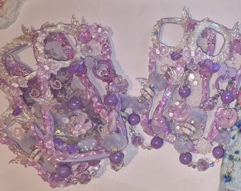 May include: Two ornate, lavender-colored phone cases. Each case is embellished with a variety of decorative elements, including beads, flowers, and dangling purple orbs. The cases have a whimsical, fairy-tale aesthetic.
