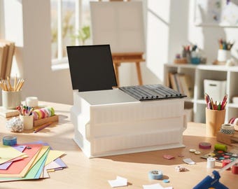 May include: A white plastic art supply organiser with a black lid is open on a wooden table. The table is covered with colourful art supplies, including paper, tape, and markers. An easel and window are in the background.
