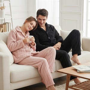 Może przedstawiać: Para w dopasowanych pluszowych piżamach, jedna w kolorze różowym, druga w czarnym, siedzi na białej kanapie. Różowa piżama składa się z bluzki z kołnierzykiem i spodni. Czarna piżama również ma bluzkę z kołnierzykiem i spodnie. Przed nimi stoi drewniany stolik kawowy.