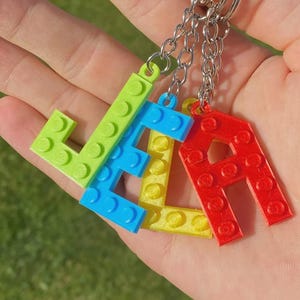 May include: A set of four letter keychains spelling the name LELA, each crafted from plastic building blocks in bright lime green, blue, yellow, and red. Each letter features a textured surface with circular studs and is attached to a silver metal ring.
