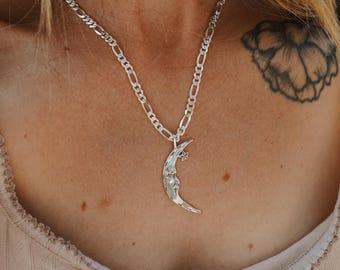 Puede incluir: Collar plateado con un colgante de media luna con una cara y una pequeña estrella. La cadena tiene un diseño de eslabones. El collar se lleva sobre una parte superior de color claro.