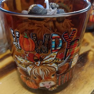 Puede incluir: Una vela decorativa en un vaso marrón con un diseño de otoño. El vaso presenta un gráfico de vaca, calabazas y la palabra "Howdy". La vela está rematada con elementos decorativos y una mecha blanca.