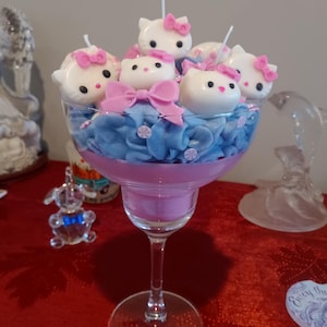 Puede incluir: Arreglo decorativo de velas en una copa de margarita. El arreglo presenta velas blancas con forma de gato con lazos rosas, colocadas sobre elementos decorativos azules y rosas. La copa está sobre una superficie roja.