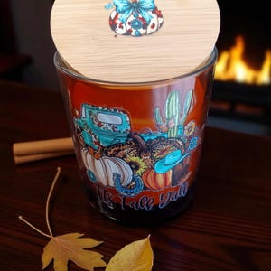 Puede incluir: Un portavelas de cristal con tapa de madera. El portavelas presenta un diseño colorido con temática otoñal con calabazas, una camioneta y el texto "It's Fall Y'all". La tapa tiene un diseño de calabaza con un lazo azul.