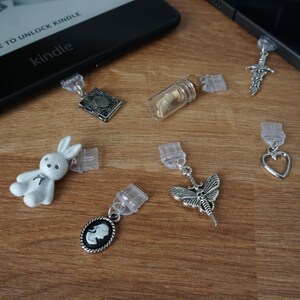 May include: A collection of decorative book page clips with various charms, including a bunny, a cameo, a moth, a sword, a heart, a small book, and a tiny glass bottle. The clips are clear, and the charms are silver-toned.