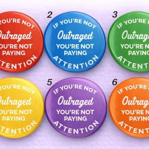 May include: A collection of six round buttons in various colors: red, blue, green, yellow, purple, and orange. Each button features the white text "IF YOU'RE NOT Outraged YOU'RE NOT PAYING ATTENTION" in a bold, sans-serif font.