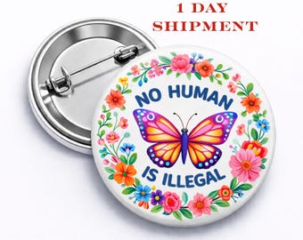 Puede incluir: Un botón redondo con un diseño de mariposa colorida y el texto "NO HUMAN IS ILLEGAL". La mariposa tiene alas rosas, amarillas y moradas. El botón está rodeado por una corona floral con flores rosas, naranjas y azules.