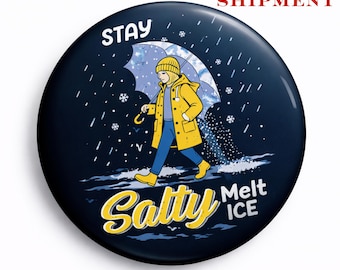 Puede incluir: Un botón redondo azul marino con una ilustración de dibujos animados de una persona con impermeable y botas amarillas caminando bajo la lluvia con un paraguas. El texto dice "STAY Salty Melt ICE".