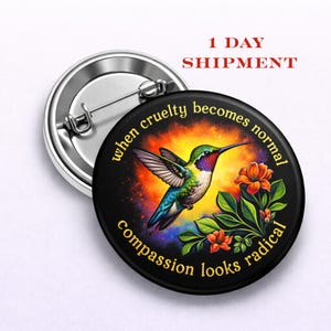 May include: A black button pin with a colorful hummingbird illustration and the text "when cruelty becomes normal, compassion looks radical." The hummingbird has green, blue, and red feathers. Orange flowers and green leaves are also featured.