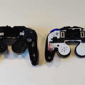 May include: Two black video game controllers, disassembled, revealing internal components. One controller is fully intact, while the other shows its circuit board and wiring. The controllers are set against a white background.