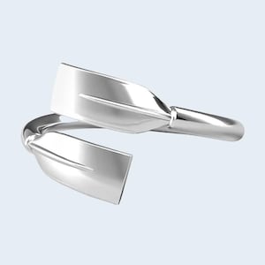 May include: A silver-toned bypass ring featuring two overlapping paddle-shaped ends. The design mimics the form of rowing oars with a smooth, reflective metallic finish, creating a continuous wrap-around band structure with an open front design.