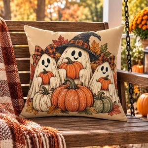 May include: A square throw pillow featuring a beige background with a printed illustration of three white ghosts wearing orange and black plaid bows and a witch hat, surrounded by orange pumpkins and colorful autumn leaves on a wooden surface.