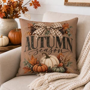 May include: A square throw pillow with a tan background featuring the text AUTUMN season in dark lettering. The design includes a large beige and white gingham bow, orange and white pumpkins, pinecones, and autumn leaves in shades of brown.