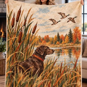 Peut inclure: Une courtepointe rectangulaire présentant une scène extérieure imprimée avec un chien brun portant un collier orange assis dans de hautes herbes de marais. L'arrière-plan montre un lac, des arbres d'automne et trois canards volant dans un ciel bleu clair.