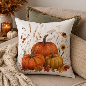 May include: A square throw pillow featuring a watercolor-style illustration of three pumpkins in shades of orange and muted green, surrounded by dried wheat stalks, wildflowers, and fallen autumn leaves on a cream-colored fabric background.