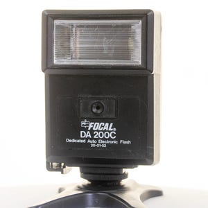 May include: A black Focal DA 200C dedicated auto electronic flash unit with a rectangular light panel at the top. The front face displays the brand name Focal, the model number DA 200C, and the text Dedicated Auto Electronic Flash 20-01-52.
