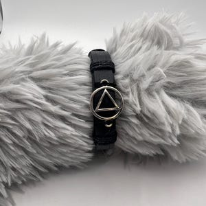 Può includere: Bracciale in pelle nera con un simbolo triangolare argentato. Il bracciale ha un design semplice con chiusura a fibbia. Il simbolo argentato è un punto focale importante, incastonato sulla pelle scura.
