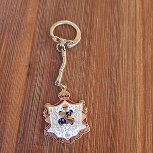 Può includere: Un portachiavi argentato con un ciondolo decorativo. Il ciondolo presenta un disegno a stemma con accenti rossi, bianchi, dorati e blu. Il portachiavi è su una superficie di legno.