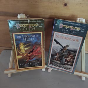 May include: Two mass-market paperback books from the Dragonlance Heroes series. The left book is Volume One, The Legend of Huma by Richard A. Knaak, featuring a yellow and red dragon. The right book is Volume Two, Stormblade by Nancy Varian Berberick.