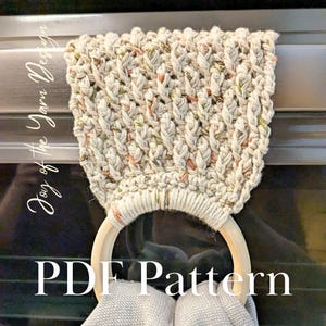 May include: A cream-colored, textured, macrame towel holder with a wooden ring. The holder is attached to a metal bar. The text "PDF Pattern" is displayed at the bottom of the image.