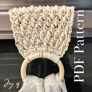 May include: Cream-colored, hand-knitted towel holder with a wooden ring. The holder features a textured, woven design with flecks of orange and green. The text "PDF Pattern" is visible on the right side. The words "Joy of The Yarn Designs" are at the bottom.