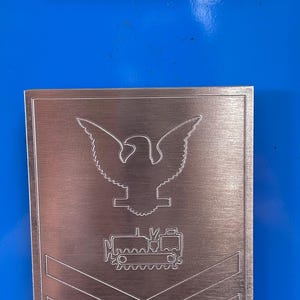 May include: Two metal plaques with military insignia. The top plaque is rectangular and features a detailed American flag design. The bottom plaque has an eagle emblem, a vehicle, and the word "ROKIS".