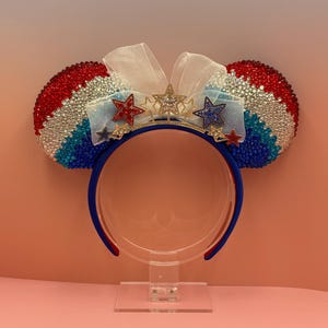 May include: A headband featuring two circular ears covered in red, white, and blue rhinestones arranged in stripes. A sheer white bow sits between the ears, adorned with five small star-shaped charms in red, white, blue, and gold tones.