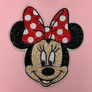 Può includere: Toppa di Minnie Mouse tempestata di strass. Il design presenta un fiocco rosso a pois bianchi, orecchie nere e un viso color oro. La toppa è posta su uno sfondo rosa.