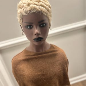 May include: A short, platinum blonde wig with tight, textured curls styled in a pixie cut. The wig is displayed on a mannequin head with dark skin, green eyes, and dark lipstick, wearing a plain, brown, short-sleeved, crew-neck top.