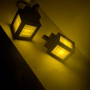 May include: Two dark, cube-shaped lanterns with a yellow glow emanating from within. The lanterns are positioned at an angle, casting a warm light in a dimly lit space. The design is reminiscent of pixel art.