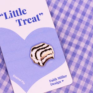 Op de afbeelding: Emaille pin met een schelpontwerp, met witte, gouden en zwarte strepen. De pin zit op een lichtblauwe kaart met de tekst "Little Treat" en "Faith Miller Design". De achtergrond is een paars en wit geruit patroon.