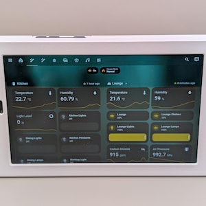 Può includere: Un pannello di controllo domestico intelligente bianco visualizza dati in tempo reale per ambienti cucina e soggiorno. Lo schermo mostra la temperatura in gradi Celsius, l'umidità, i livelli di luce e le opzioni di controllo per vari dispositivi. L'interfaccia è intuitiva.