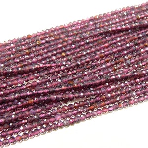 Könnte beinhalten: Nahaufnahme einer Sammlung kleiner, facettierter, tiefrot-violetter Edelsteinperlen. Die Perlen sind aufgefädelt und bilden eine dichte, strukturierte Oberfläche. Die Farbe ist durchgehend, mit subtilen Farbtonvariationen.