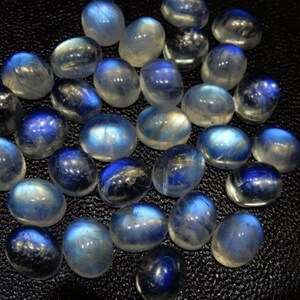 K&ouml;nnte beinhalten: Eine Sammlung polierter Mondstein-Cabochons. Die Steine zeigen eine Farbpalette von durchscheinendem Wei&szlig; bis tiefem Blau mit einem schimmernden, irisierenden Effekt. Die oval geformten Steine sind auf einer dunklen Oberfl&auml;che angeordnet.
