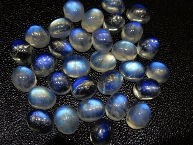 K&ouml;nnte beinhalten: Eine Sammlung polierter Mondstein-Cabochons. Die Steine zeigen eine Farbpalette von durchscheinendem Wei&szlig; bis tiefem Blau mit einem schimmernden, irisierenden Effekt. Die oval geformten Steine sind auf einer dunklen Oberfl&auml;che angeordnet.