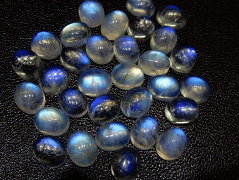 K&ouml;nnte beinhalten: Eine Sammlung polierter Mondstein-Cabochons. Die Steine zeigen eine Farbpalette von durchscheinendem Wei&szlig; bis tiefem Blau mit einem schimmernden, irisierenden Effekt. Die oval geformten Steine sind auf einer dunklen Oberfl&auml;che angeordnet.