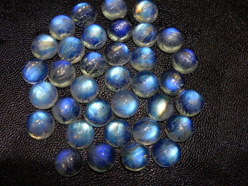 K&ouml;nnte beinhalten: Eine Sammlung runder, polierter Mondstein-Cabochons. Die Steine zeigen eine Reihe von Blau- und Wei&szlig;t&ouml;nen mit einer schimmernden, irisierenden Qualit&auml;t. Die Cabochons sind auf einer dunklen, strukturierten Oberfl&auml;che angeordnet.