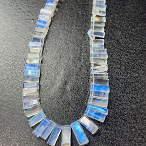 May include: A necklace composed of rectangular, translucent moonstone beads. The beads have a subtle blue iridescence, strung together to form a curved shape. The necklace is displayed against a dark, textured background.