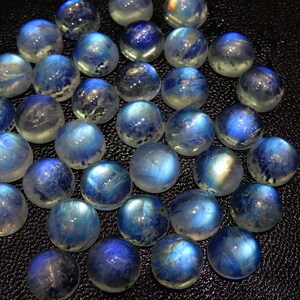 K&ouml;nnte beinhalten: Eine Ansammlung runder, polierter Mondstein-Cabochons. Die Steine zeigen eine Bandbreite an Blau- und Wei&szlig;t&ouml;nen mit einer schimmernden, irisierenden Qualit&auml;t. Die Cabochons sind eng angeordnet und zeigen ihre glatten, gew&ouml;lbten Oberfl&auml;chen.