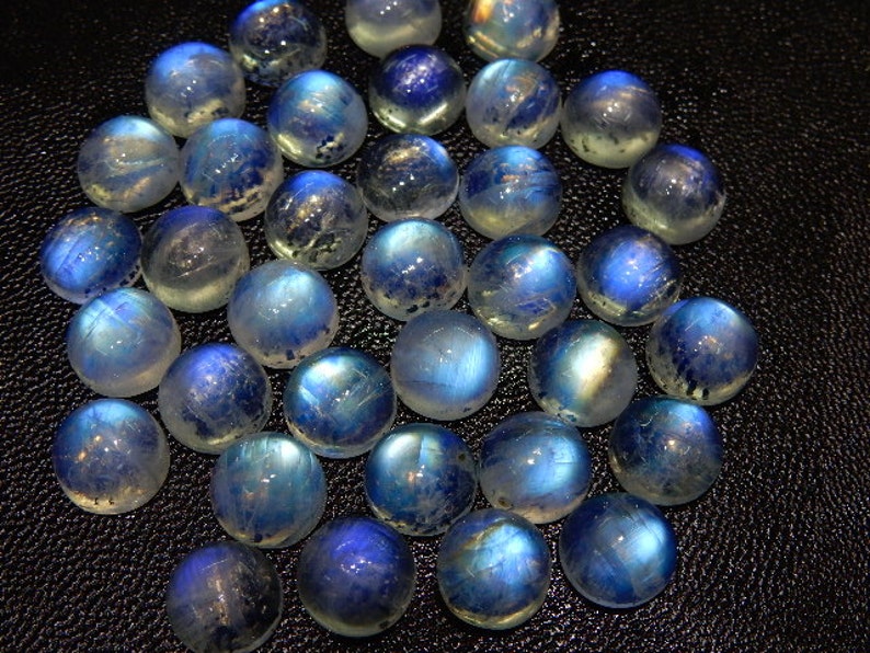 K&ouml;nnte beinhalten: Eine Ansammlung runder, polierter Mondstein-Cabochons. Die Steine zeigen eine Bandbreite an Blau- und Wei&szlig;t&ouml;nen mit einer schimmernden, irisierenden Qualit&auml;t. Die Cabochons sind eng angeordnet und zeigen ihre glatten, gew&ouml;lbten Oberfl&auml;chen.