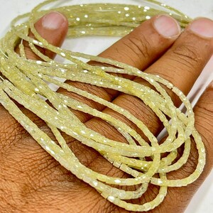 May include: A hand holds strands of pale yellow, square-cut beads with a slight shimmer. The beads are looped, creating a layered effect. The overall colour scheme is soft and natural. The image has a date stamp.