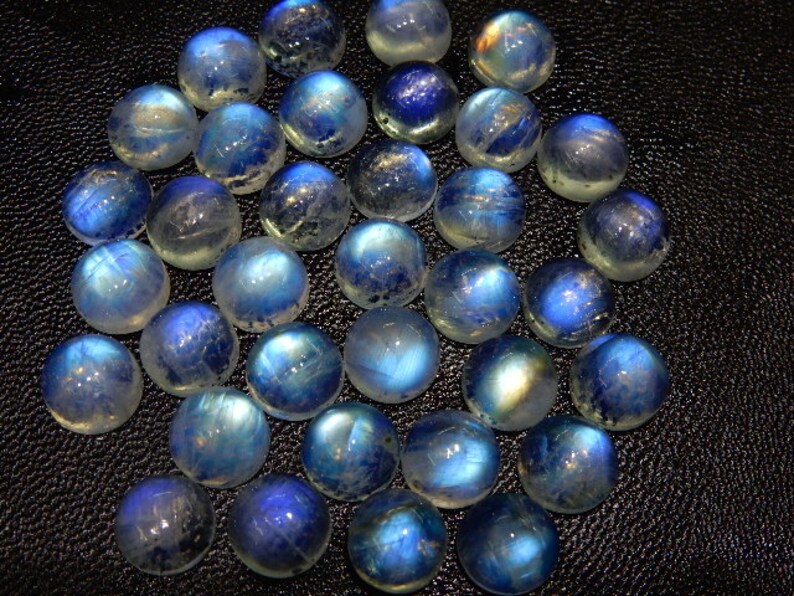 K&ouml;nnte beinhalten: Eine Sammlung runder, polierter Mondstein-Cabochons. Die Steine zeigen eine Mischung aus blauen und wei&szlig;en Farben mit einem durchscheinenden Aussehen. Die Edelsteine sind eng aneinander angeordnet und zeigen ihre nat&uuml;rliche Sch&ouml;nheit und ihr Potenzial f&uuml;r die Schmuckherstellung.