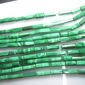 Puede incluir: Primer plano de hilos de cuentas de malaquita. Las cuentas son rectangulares con un acabado pulido, que muestra un color verde vibrante con patrones arremolinados de color verde más oscuro. Las cuentas están ensartadas, listas para la fabricación de joyas.