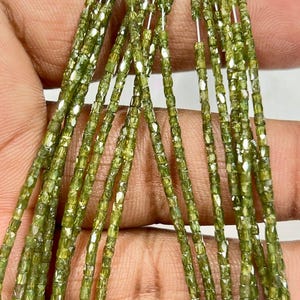 May include: Close-up of multiple strands of small, olive-green beads. The beads are rectangular and faceted, catching the light. The overall effect is of a delicate, shimmering material. The beads are likely gemstones.