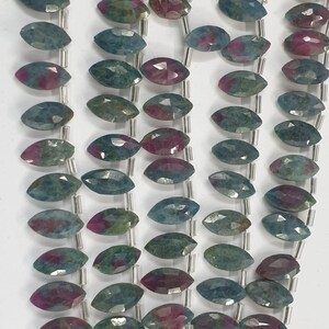 Puede incluir: Primer plano de hileras de cuentas de piedras preciosas facetadas. Las cuentas ovaladas presentan tonos de verde azulado y magenta. Están ensartadas en un alambre plateado. Las facetas de las piedras preciosas reflejan la luz.