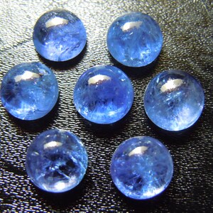 May include: Seven round, polished blue gemstones with a translucent appearance are arranged on a dark surface. The stones have a vibrant, deep blue color with internal patterns and a glossy finish, reflecting light.