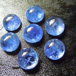 May include: Seven round, translucent blue gemstones are arranged on a dark surface. The stones have a polished, glassy appearance, with internal textures and variations in color. The gems are illuminated, creating highlights and shadows that enhance their three-dimensional form.
